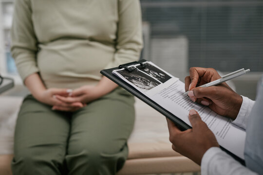 Pregnant woman sitting on examination table during prenatal checkup while doctor holding clipboard with ultrasound images and filling out medical form in clinic setting - Powered by Adobe