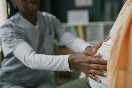 Black female healthcare professional examining pregnant Asian woman during prenatal care checkup, hands gently touching abdomen, medical consultation in clinical setting