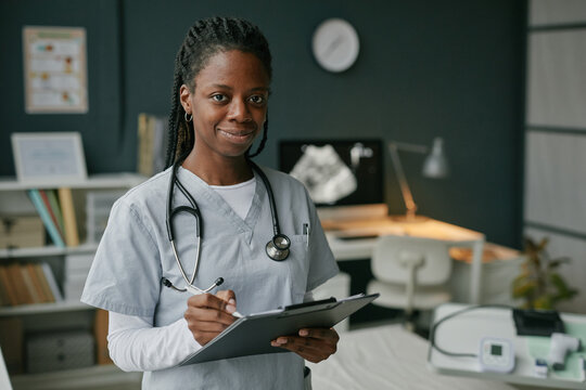 Portrait of young adult Black woman doctor standing in medical office holding clipboard and pen smiling at camera providing prenatal care checkup - Powered by Adobe