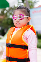 Cute little Asian girl wearing life jacket playing in swimming pool,Asian girl wearing life jacket sits on edge of pool and plays in water in swimming pool, summer leisure activity.