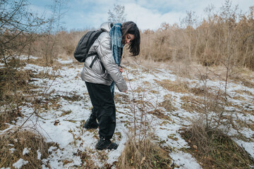 A young woman dressed in winter attire is hiking through a partially snow-covered forested area...
