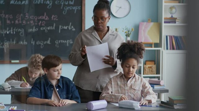 Medium shot of African American female teacher handing out worksheets to diverse students during classroom activity