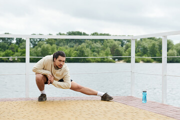 Sporty man stretches by the waterfront on a cloudy afternoon while wearing a hoodie