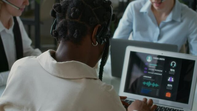 Rack focus view of African American young female call centre employee clarifying customers issue while working at helpline using laptop at table with other colleagues