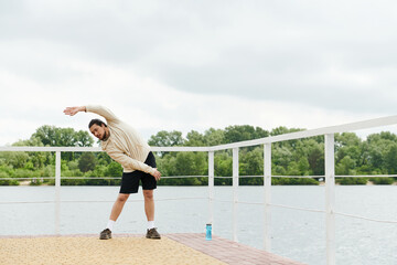 Sporty man stretching outdoors by the water on a cloudy day
