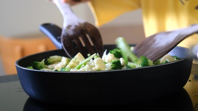 A person stirs fresh vegetables like broccoli, cauliflower, and celery in a black non-stick pan using wooden utensils, highlighting healthy home-cooked meal preparation.