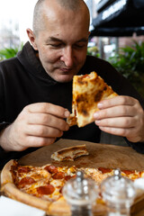 A man with a glass of dark beer in his hand enjoys a slice of pizza. There is a salt and pepper shaker and a kettle on the table.