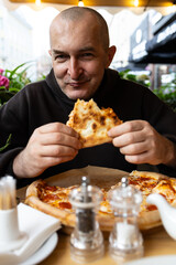 A man with a glass of dark beer in his hand enjoys a slice of pizza. There is a salt and pepper shaker and a kettle on the table.