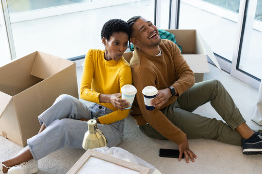Leaning diverse couple unpacking boxes on living room floor, holding takeaway coffee cups