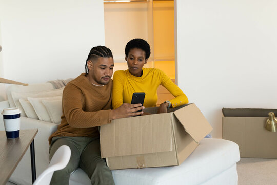 Diverse couple checking smartphone sitting on beige sofa in living room, with cardboard boxes