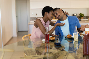 Laughing African American couple feeding each other burgers at kitchen dining table, with drinks