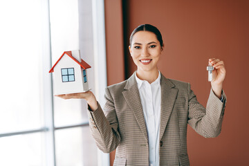 Confident businesswoman holding house model and keys, symbolizing real estate success and property...
