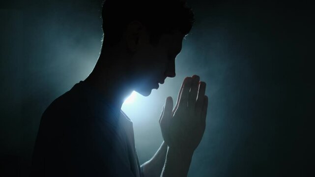 Silhouette of desperate man praying in dim blue light, expressing emotional breakdown and spiritual seeking in solitude.