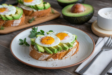 Healthy breakfast plate with toasted sourdough, sliced avocado and two sunny side up eggs surrounded by whole and halved avocados on rustic table