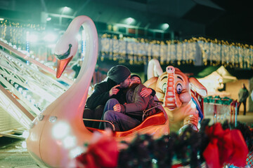 A couple seated in a swan-shaped ride at a brightly lit outdoor carnival decorated with lights and...