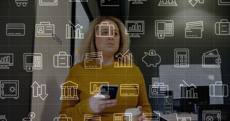Holding smartphone and coffee, woman in mustard sweater looking up in office with finance icons