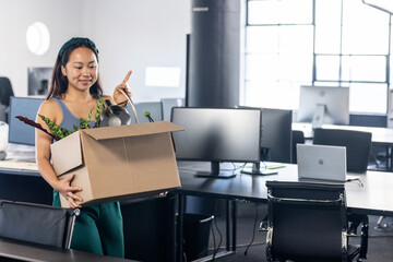 Chinese woman walking through office with moving box containing potted plant, desk lamp, copy space