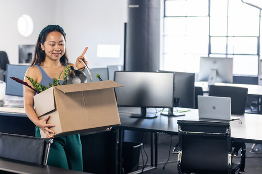 Chinese woman walking through office with moving box containing potted plant, desk lamp, copy space