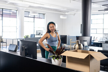 Asian Chinese woman standing unpacking items at open-plan office desk with monitor, plant and box