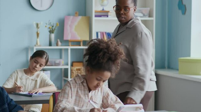 Medium shot of helpful African American female teacher guiding diverse group of students concentrated on classwork at their desks, highlighting contribution of teacher in education process