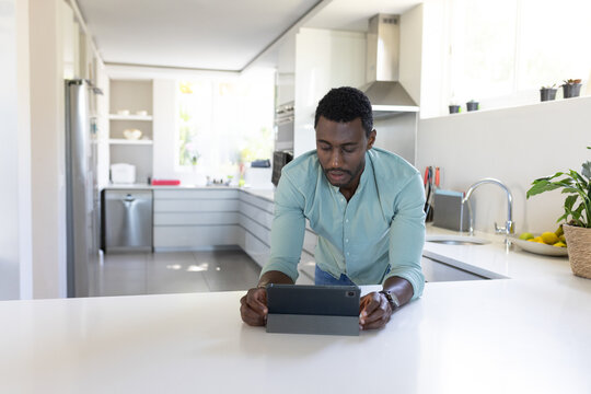 African American man leaning on kitchen island counter, using tablet computer near potted plant