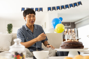 Asian man sitting on sofa marking birthday with cake, balloons, birthday banner, copy space