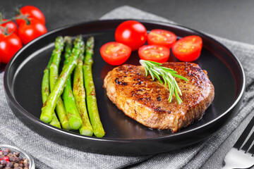 Close up, macro of tasty grilled beef steak served with green asparagus and cherry tomatoes on black plate, with cutlery. Perfect for food menu, diet, or culinary design concepts.