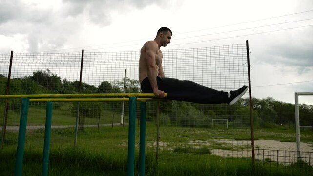 Strong man trains abdominal muscles raising legs on parallel bars at sports ground. Young shirtless sportsman exercising at nature. Sporty athlete working out at nature. Concept of sportive lifestyle