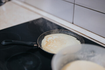 A fresh pancake being cooked on a non-stick frying pan atop a hot stove. Steam rises, depicting the cooking process, accompanied by a bowl of batter in the foreground.