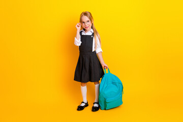 Smiling young schoolgirl in uniform with backpack standing against bright yellow background ready...