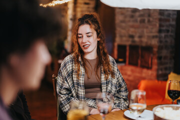 A young woman enjoys a friendly conversation during a relaxed dinner gathering, with drinks and snacks on the table. The warm indoor ambiance complements the joyful atmosphere of the evening.