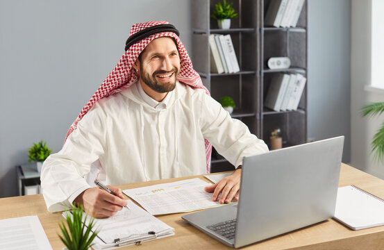Happy Arabian man working on financial report, writing in paper documents and using laptop for work, wearing traditional ghutra on head, has cheerful expression, posing against office interior