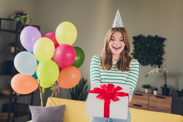 A joyful young woman celebrating with balloons and a gift in a cozy home interior, showcasing...
