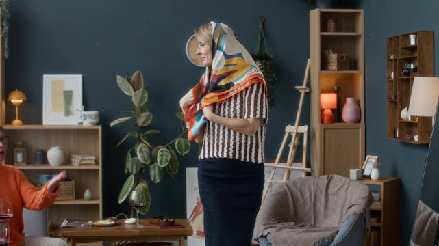 Mature Caucasian woman trying on babushka scarf in front of her best friends in modern living room
