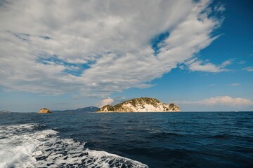 Waves gently lap against a small, rocky island surrounded by sparkling blue waters. Fluffy clouds...