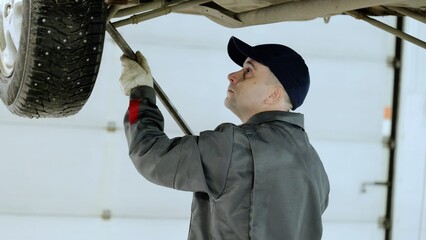 Professional mechanic repairing vehicle, lifting heavy automotive part with crowbar, wearing...