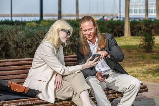 Business colleagues, Albino woman and redhead Man, looking at Smartphone on a bench in a city Park