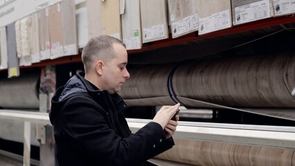 Male customer focusing on smartphone while comparing rolled vinyl flooring, calculating prices and...