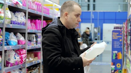 Focused shopper examining detergent bottle label, comparing cleaning products amid supermarket...