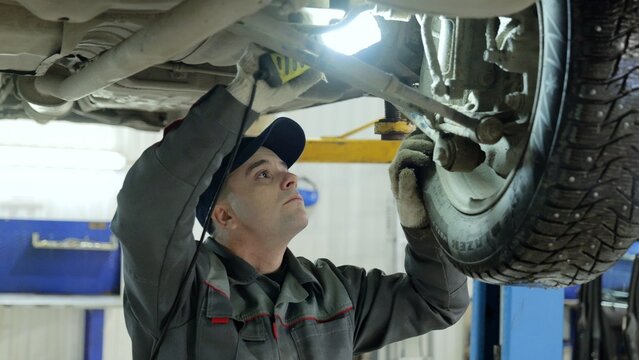 Skilled mechanic examining vehicle underside with bright flashlight, wearing protective gloves inside modern automotive workshop during detailed technical inspection