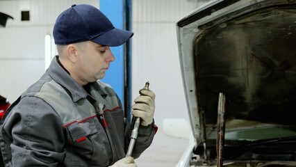 Auto mechanic wearing protective gloves, examining car engine in workshop, holding spark plug and...