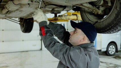 Experienced automotive technician checking vehicle undercarriage, wearing protective gear and using...