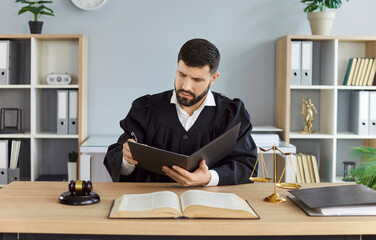 Portrait of confident young man judge wearing judicial robe sitting at the desk with gavel and...