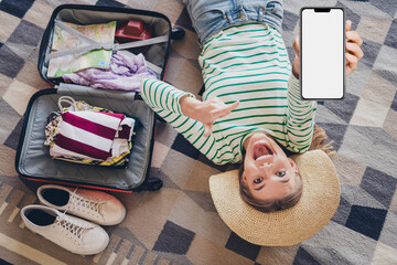 Happy young woman smiling and showing smartphone while packing suitcase at home for a vacation