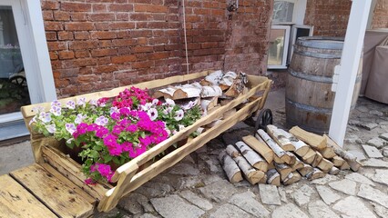 Rustic decor in courtyard of house. Wooden rural cart with birch firewood and bright fresh flowers....