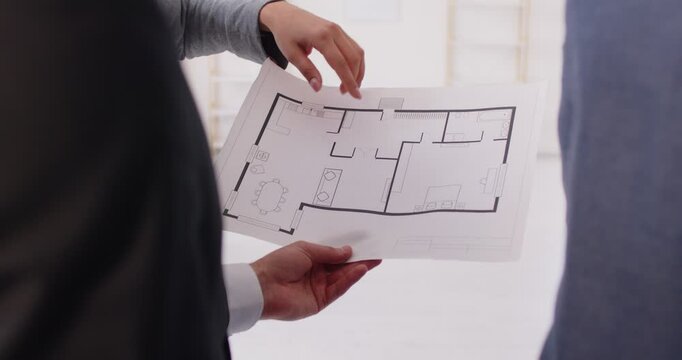 Hands holding document displaying floor plan close up, pointing to section on paper, examining layout discussing details about space on blueprint, real estate agent explaining or presenting to clients