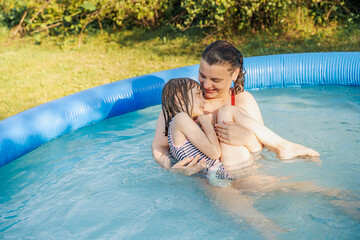 Mother lifts daughter in inflatable pool on sunny summer day. Girl laughing, splashing water....
