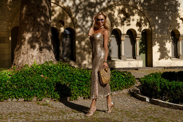 Elegant female model in gold sequin mini dress and fashionable boots posing in the city in summer...