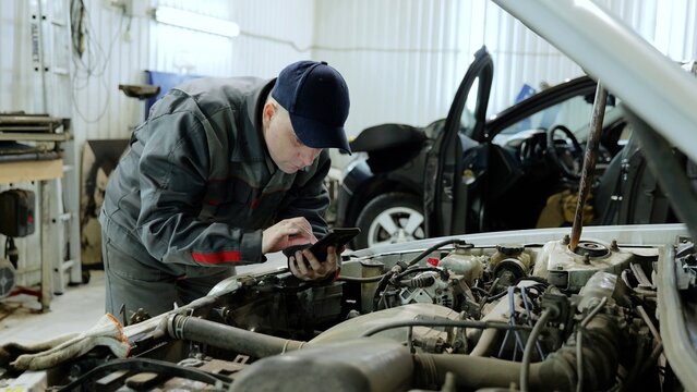 Professional mechanic using a mobile phone for diagnostics while inspecting a vehicle engine in a garage, showcasing modern automotive repair technology