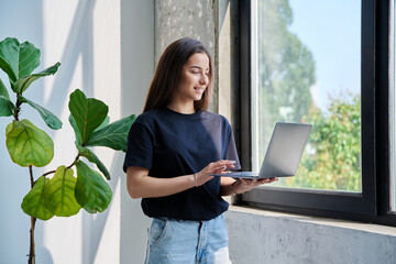 Portrait of young woman standing with laptop computer in home interior near window